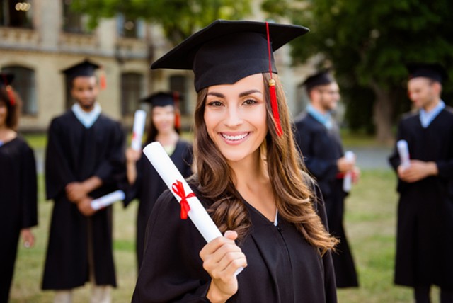 Happy cute brunette Caucasian grad girl is smiling, blurred classmates are behind. She is in a black mortar board, with a red tassel, in a gown, with nice brown curly hair, diploma in hand, and waiting for Black Car Service